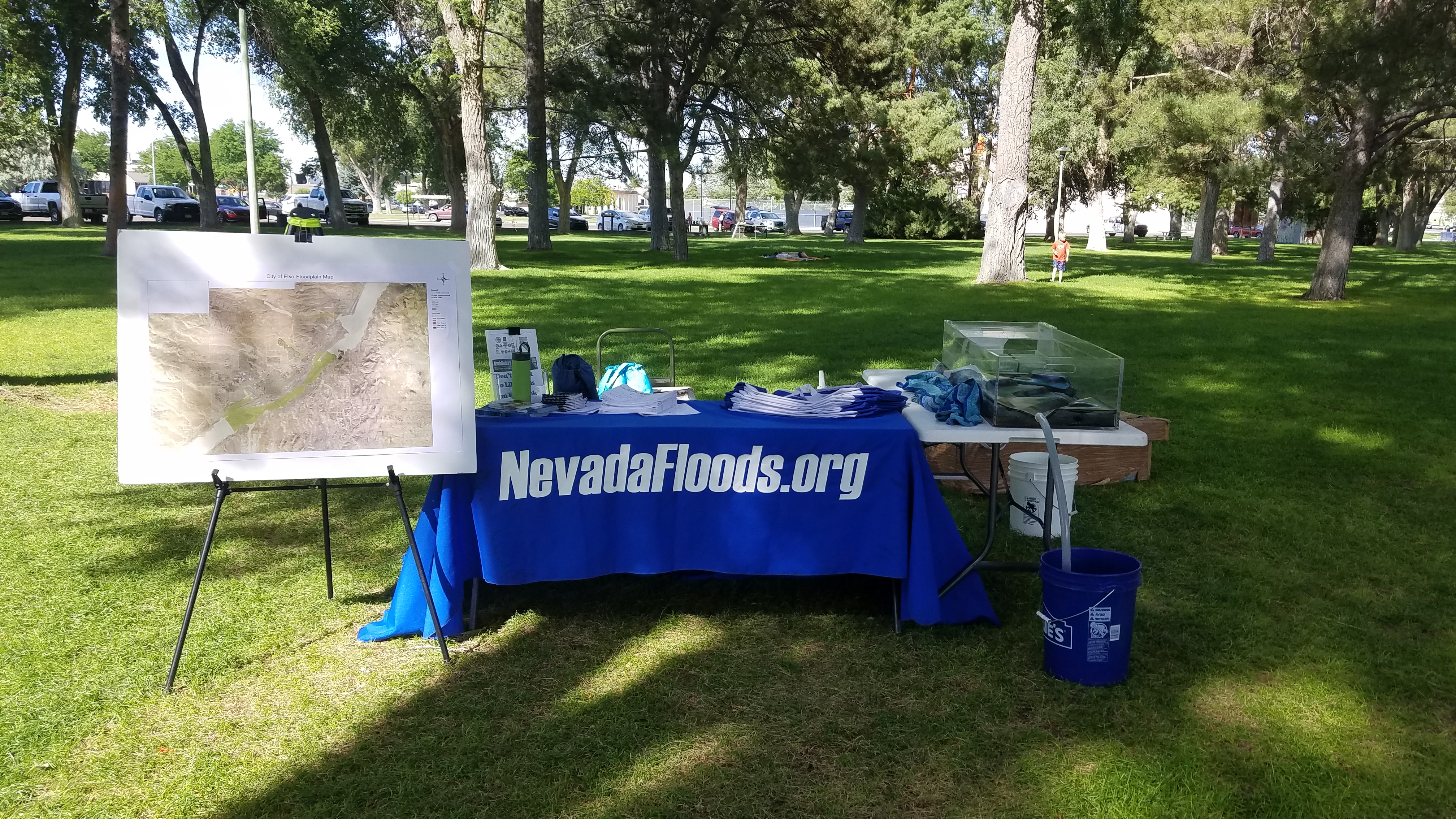 A photo image of the Nevada Floods outreach booth before an event. The booth consists of a blue table cloth that reads Nevadafloods.org and has outreach materials resting on it. The flooding simulation model is sitting on an adjacent table.